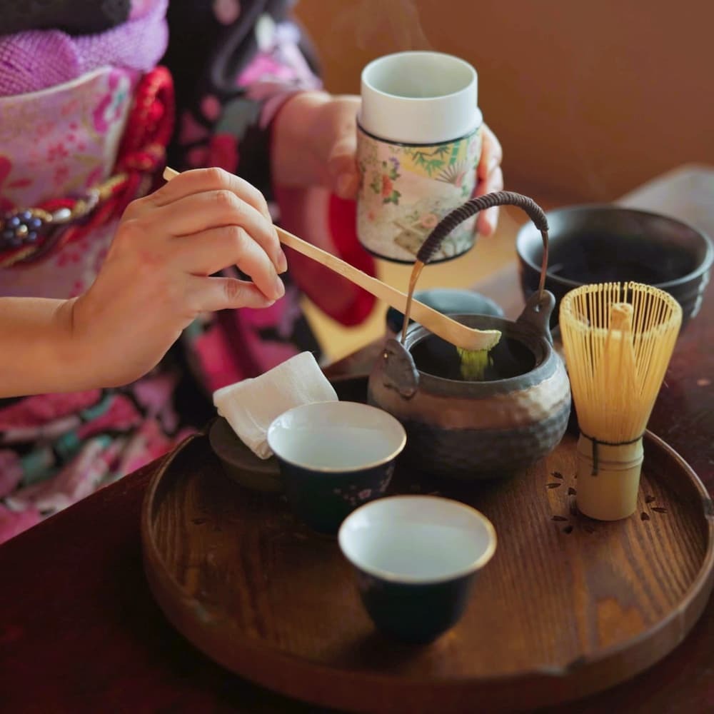 Japansk kvinna som förbereder matcha-te i en traditionell teceremoni hemma, med matchapulver, örter och en tekanna