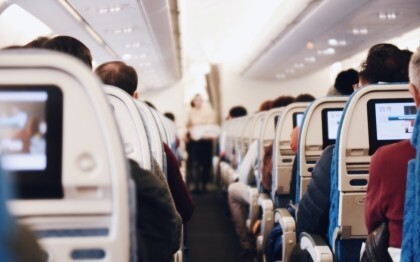 Interiör i ett stort passagerarflygplan med sittande människor och en flygvärdinna i uniform som går längs gången